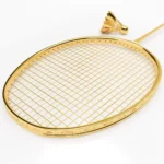 The gold head and string bed of the 24K Gold Badminton Set with the matching gold shuttlecock behind it