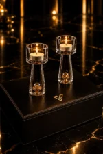 24K Gold Tealight Holders lit as a pair on a black leather box, each flame above a clear glass column with a gold leaf cluster sealed in the base